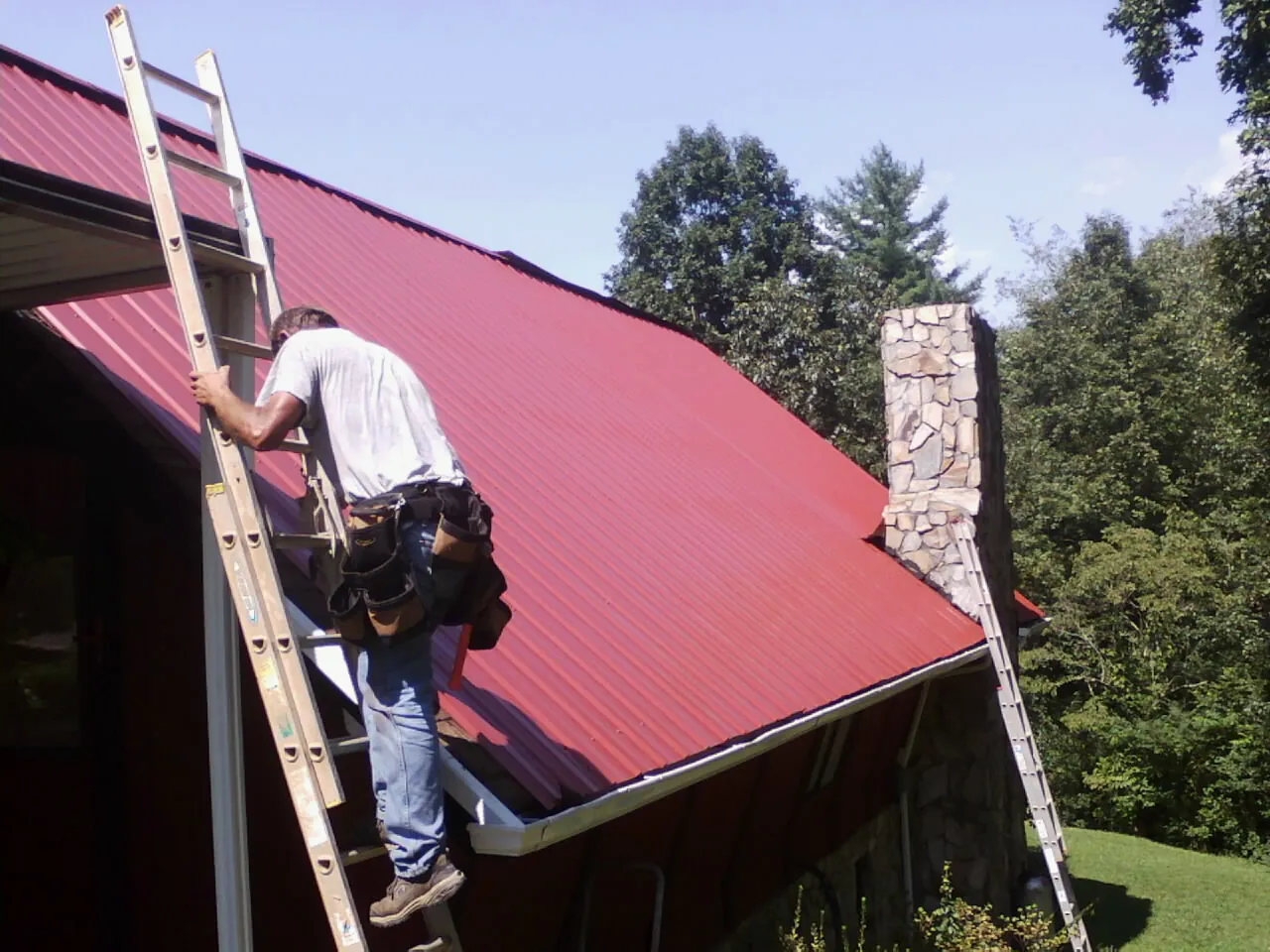Roofer on a ladder beside a red metal roof and stone chimney — Soffit & Fascia Repair by Roofing Brothers in Chimney Rock